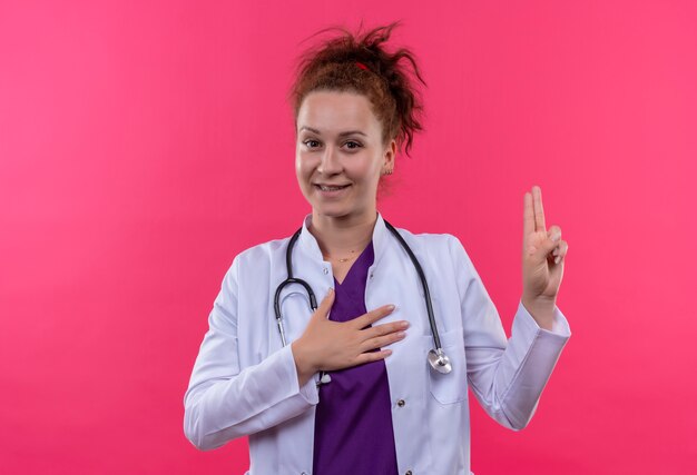 Молодая женщина-врач в белом халате со стетоскопом улыбается, принимая присягу, стоя над розовой стеной