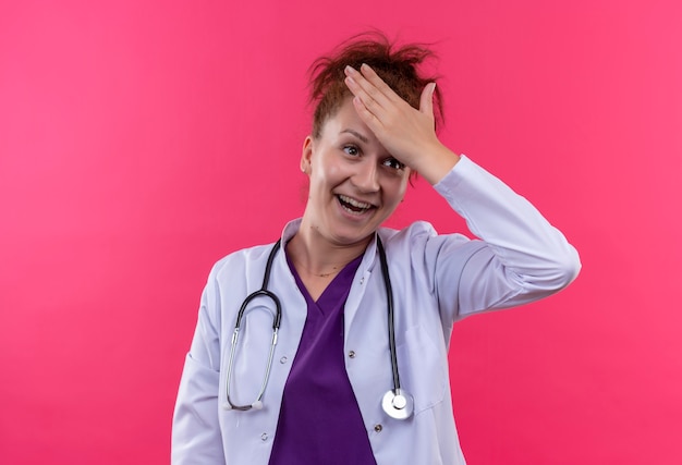 Young woman doctor wearing white coat with stethoscope smiling touching head for mistake with confuse expression on face standing over pink wall