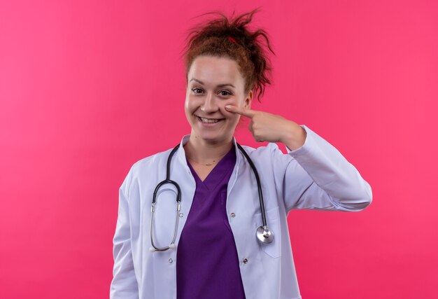 Молодая женщина-врач в белом халате со стетоскопом улыбается счастливым лицом, указывая пальцем на ее щеку, стоя над розовой стеной