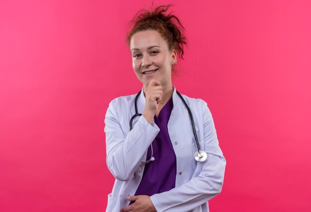 Young woman doctor wearing white coat with stethoscope  with hand on chin smiling standing over pink wall