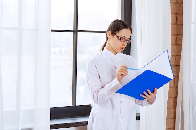 Young woman doctor in white coat reading about next patient near the window. 