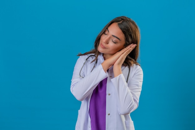 Young woman doctor in white coat with phonendoscope dreaming and posing with hands together while smiling with closed eyes over isolated blue background