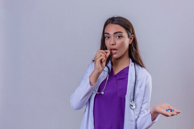 Молодая женщина-врач в белом халате с фонендоскопом, держа в руке маленькие таблетки и собирается взять один из них на белом фоне изолированные с копией пространства