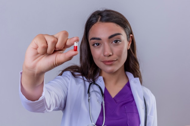 Medico della giovane donna in camice con phonendoscope che esamina macchina fotografica con il sorriso sul fronte che giudica la piccola pillola a disposizione sopra bianco ha isolato il fondo