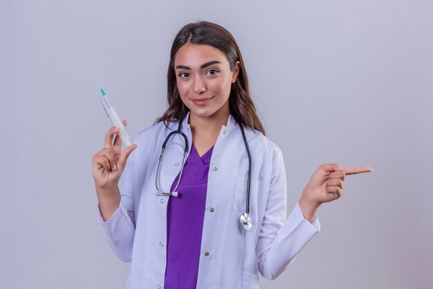Young woman doctor in white coat with phonendoscope looking confident holding syringe with medical vaccine and pointing with hand and finger to the side over isolated white background