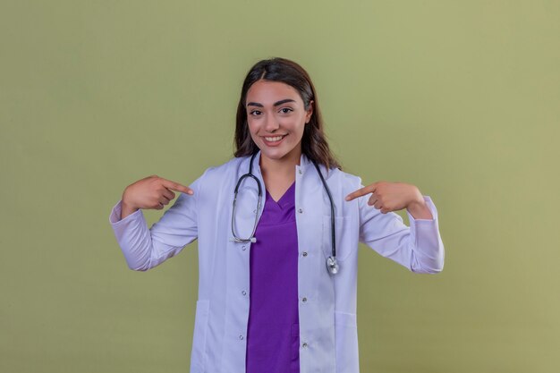 Молодая женщина-врач в белом халате с фонендоскопом, уверенно смотрит с улыбкой на лице, указывая на себя пальцами, гордыми и счастливыми, стоя над изолированным зеленым фоном