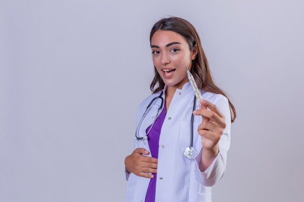 Phonendoscope 흰색 배경 위에 온도계를 보여주는 얼굴에 미소로 자신감을 찾고 흰색 코트에 젊은 여자 의사