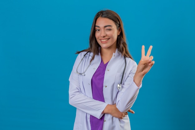 Young woman doctor in white coat with phonendoscope showing and pointing up with fingers number two or victory sign while smiling confident and happy standing over isolated blue background