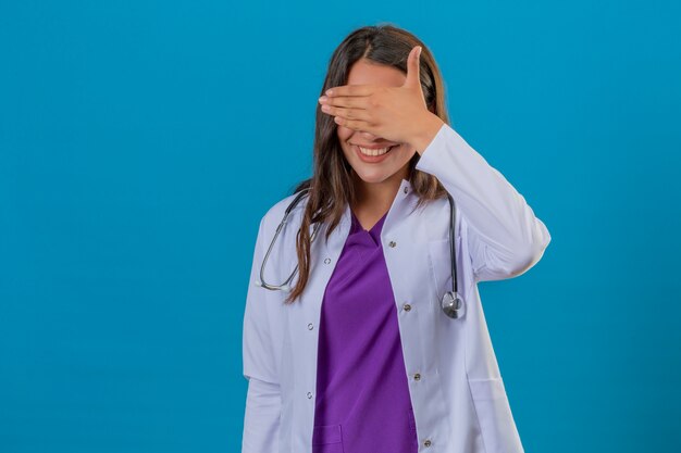 Young woman doctor in white coat with phonendoscope smiling and covering eyes by hand standing over blue background