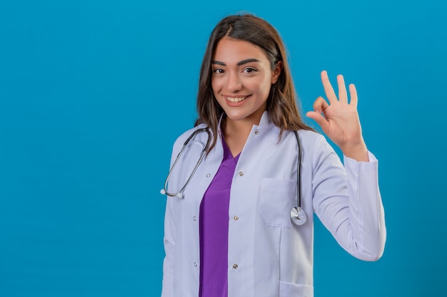 Young woman doctor in white coat with phonendoscope smiling doing ok sign with fingers over isolated blue background