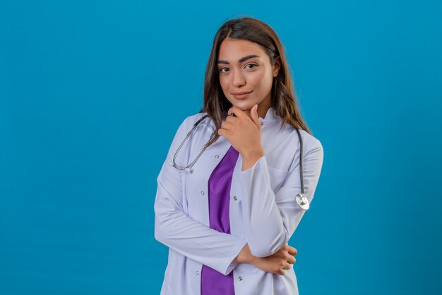 Young woman doctor in white coat with phonendoscope smiling happy and confident touching chin with hand over isolated blue background