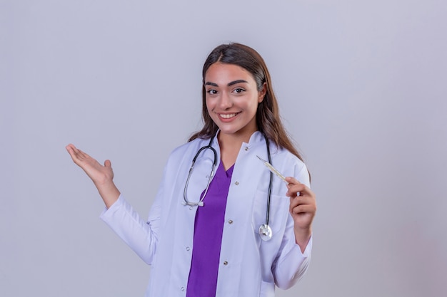 Young woman doctor in white coat with phonendoscope smiling holding thermometer and with arm out in a welcoming gesture over white isolated background