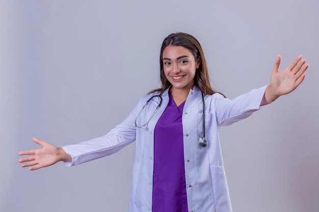 Young woman doctor in white coat with phonendoscope smiling making a welcome gesture with raised hands over white isolated background