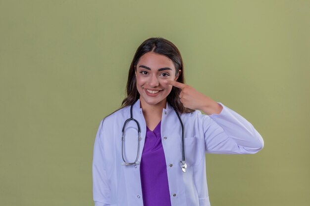 Young woman doctor in white coat with phonendoscope smiling and pointing with hand finger to face and nose standing over green isolated background