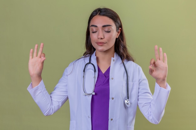Young woman doctor in white coat with phonendoscope smiling with eyes closed doing meditation gesture with fingers over green isolated background