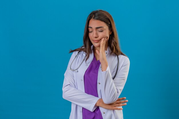 Young woman doctor in white coat with phonendoscope suffering from toothache standing over isolated blue background