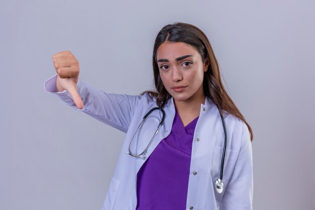 Молодая женщина-врач в белом халате с фонендоскопом с несчастным лицом, показывая неприязнь с большими пальцами руки вниз, стоя над изолированным белым фоном