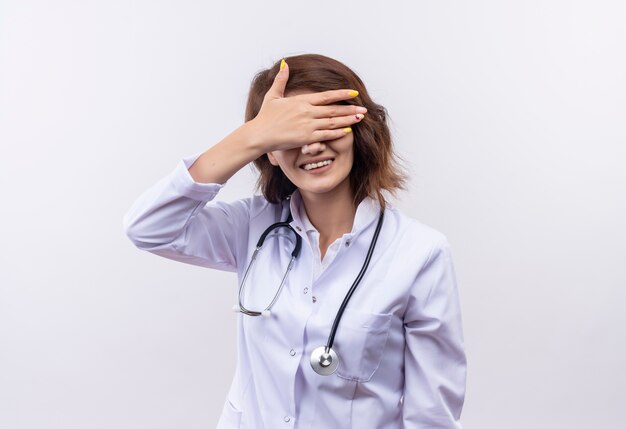 陽気な笑顔の手で目を覆う聴診器と白衣を着た若い女性医師