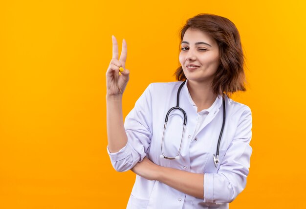 청진 기 행복 하 고 긍정적 인 윙크 승리 기호를 보여주는 흰색 코트에 젊은 여자 의사