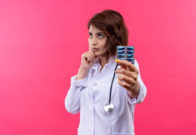 Молодая женщина-врач в белом халате со стетоскопом держит блистер с таблетками, глядя в сторону с задумчивым выражением лица