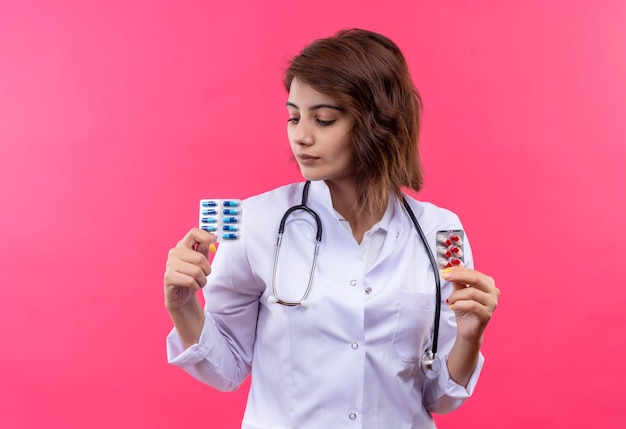 Free photo young woman doctor in white coat with stethoscope holding blister with pills looking at it with serious face