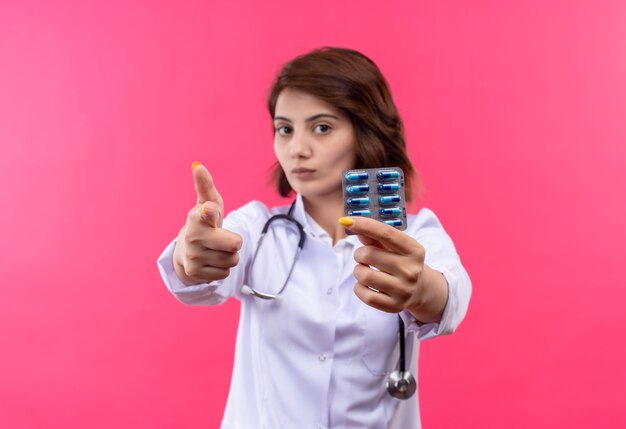 Young woman doctor in white coat with stethoscope holding blister with pills pointing with finger to camera with serious face 