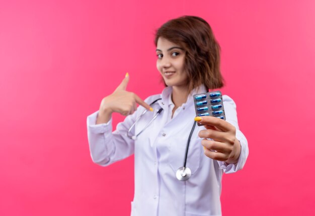 Young woman doctor in white coat with stethoscope holding blister with pills smiling confident pointing to herself 