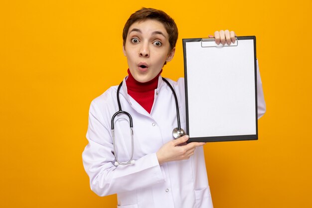 Young woman doctor in white coat with stethoscope holding clipboard with blank pages looking amazed and surprised