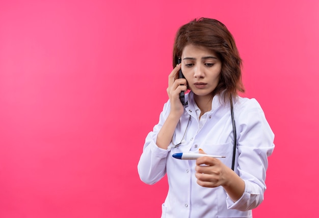 Young woman doctor in white coat with stethoscope holding digital thermometer looking at it worried while talking on mobile phone 