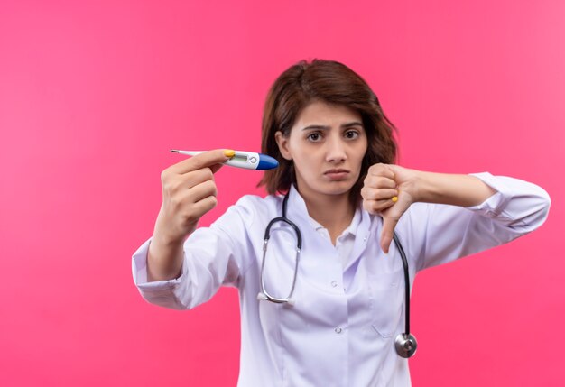 Young woman doctor in white coat with stethoscope holding digital thermometer showing thumbs down looking displeased 