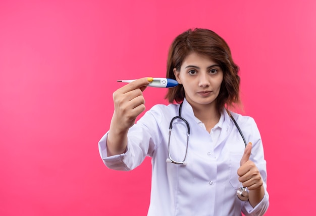 Young woman doctor in white coat with stethoscope holding digital thermometer showing thumbs up 
