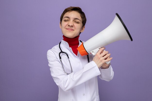 Young woman doctor in white coat with stethoscope holding megaphone smiling happy and cheerful