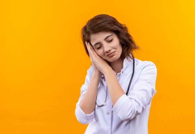 Young woman doctor in white coat with stethoscope holding palms together leaning head on palms wants to sleep