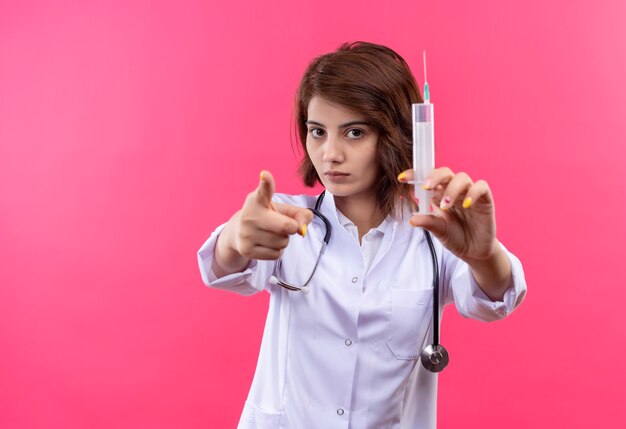 Young woman doctor in white coat with stethoscope holding syringe pointing with finger to camera 