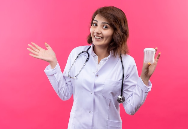 Young woman doctor in white coat with stethoscope holding test jar looking confused smiling spreading arm to the side 