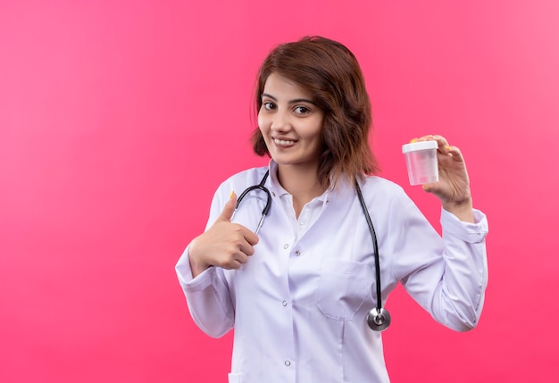 Young woman doctor in white coat with stethoscope holding test jar smiling cheerfully showing thumbs up 