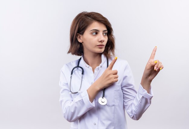 Молодая женщина-врач в белом халате со стетоскопом смотрит в сторону, уверенно указывая пальцами вверх