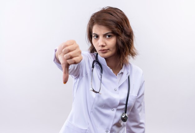 Young woman doctor in white coat with stethoscope looking at camera displeased showing thumbs down 