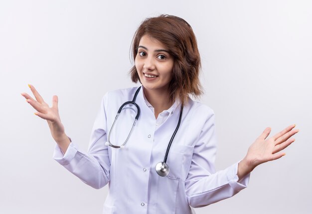 Young woman doctor in white coat with stethoscope looking at camera smiling cheerfully spreading arms to the sides 