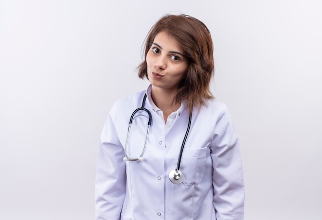 Young woman doctor in white coat with stethoscope looking at camera with shy smile on face 