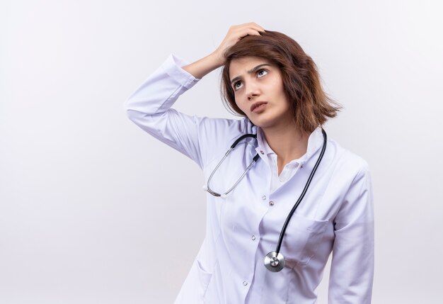 Young woman doctor in white coat with stethoscope looking confused with hand on head with puzzled 