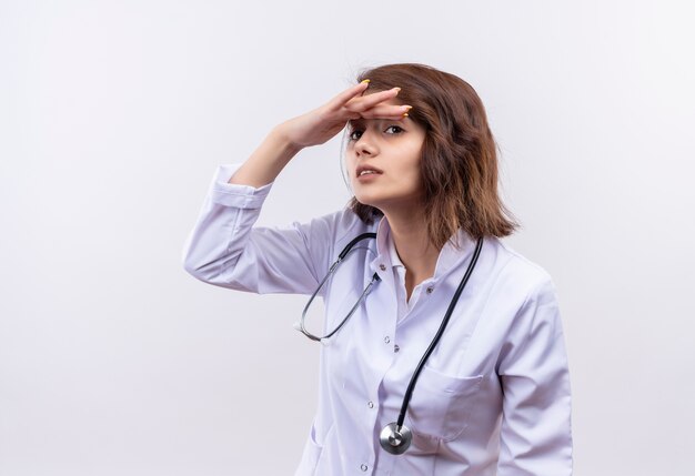 Young woman doctor in white coat with stethoscope looking faraway wit hand over head to look someone or something 