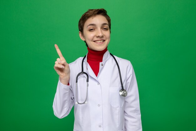 Молодая женщина-врач в белом халате со стетоскопом выглядит счастливой и позитивной, показывая указательный палец, имеющий хорошую идею