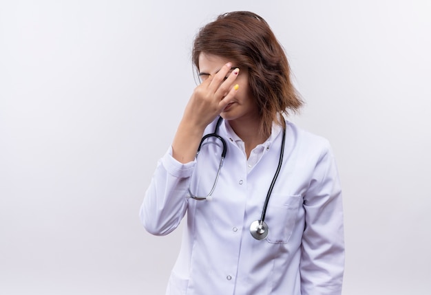 Young woman doctor in white coat with stethoscope looking tire and overworked touching nose between closed eyes 