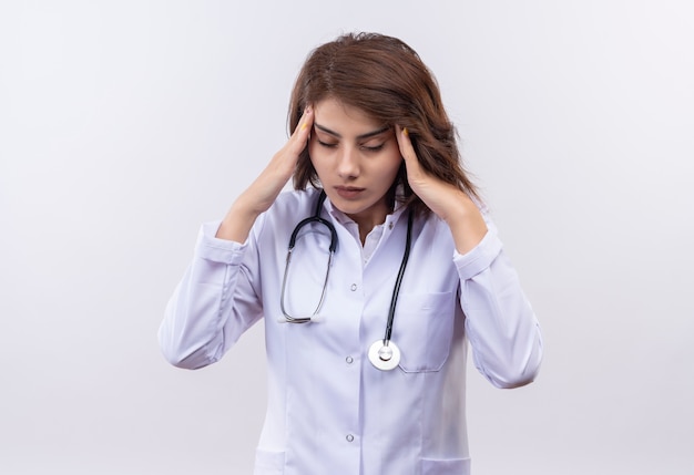 Young woman doctor in white coat with stethoscope looking tired and overworked having strong headache 