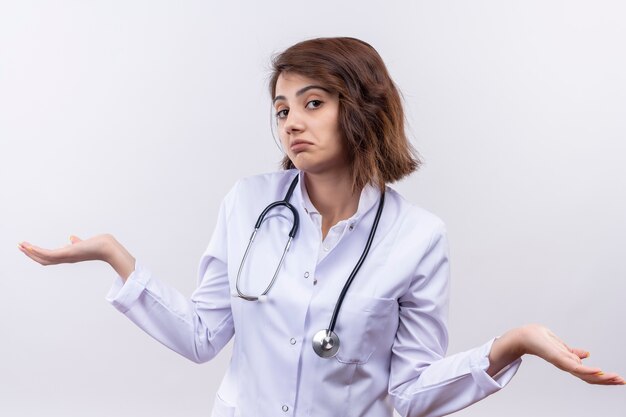 Young woman doctor in white coat with stethoscope looking uncertain and confused shrugging shoulders having no answer 