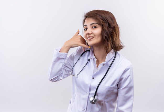 Young woman doctor in white coat with stethoscope making call me gesture smiling cheerfully standing over white wall