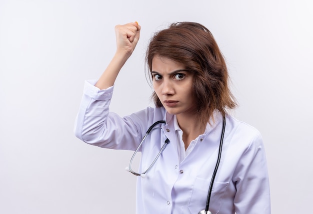 Young woman doctor in white coat with stethoscope raising fist  with angry face standing over white wall