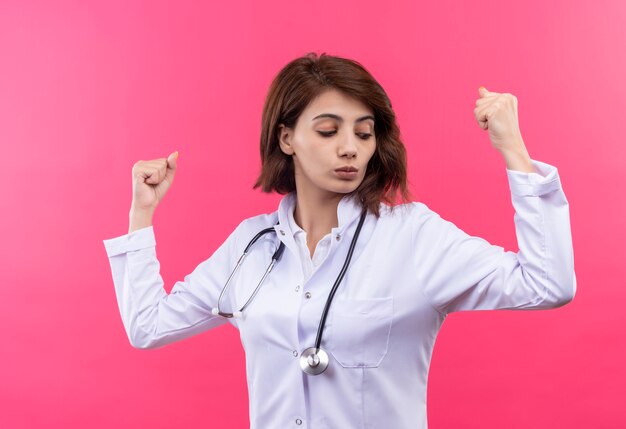 Young woman doctor in white coat with stethoscope raising fists showing biceps like winner standing over pink wall