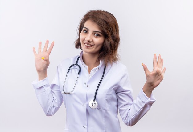Молодая женщина-врач в белом халате со стетоскопом, поднимая руки в капитуляции, улыбаясь, стоя над белой стеной